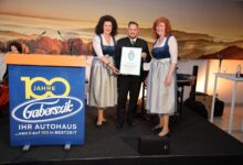 Drei Personen in bayerischer Tracht stehen auf einer Bühne bei den Holz steirischen Tischlertagen; der Mann in der Mitte hält eine Urkunde. Ein Podium mit der Aufschrift "100 Jahre Gaberszik" und ein landschaftlicher Hintergrund sind zu sehen.