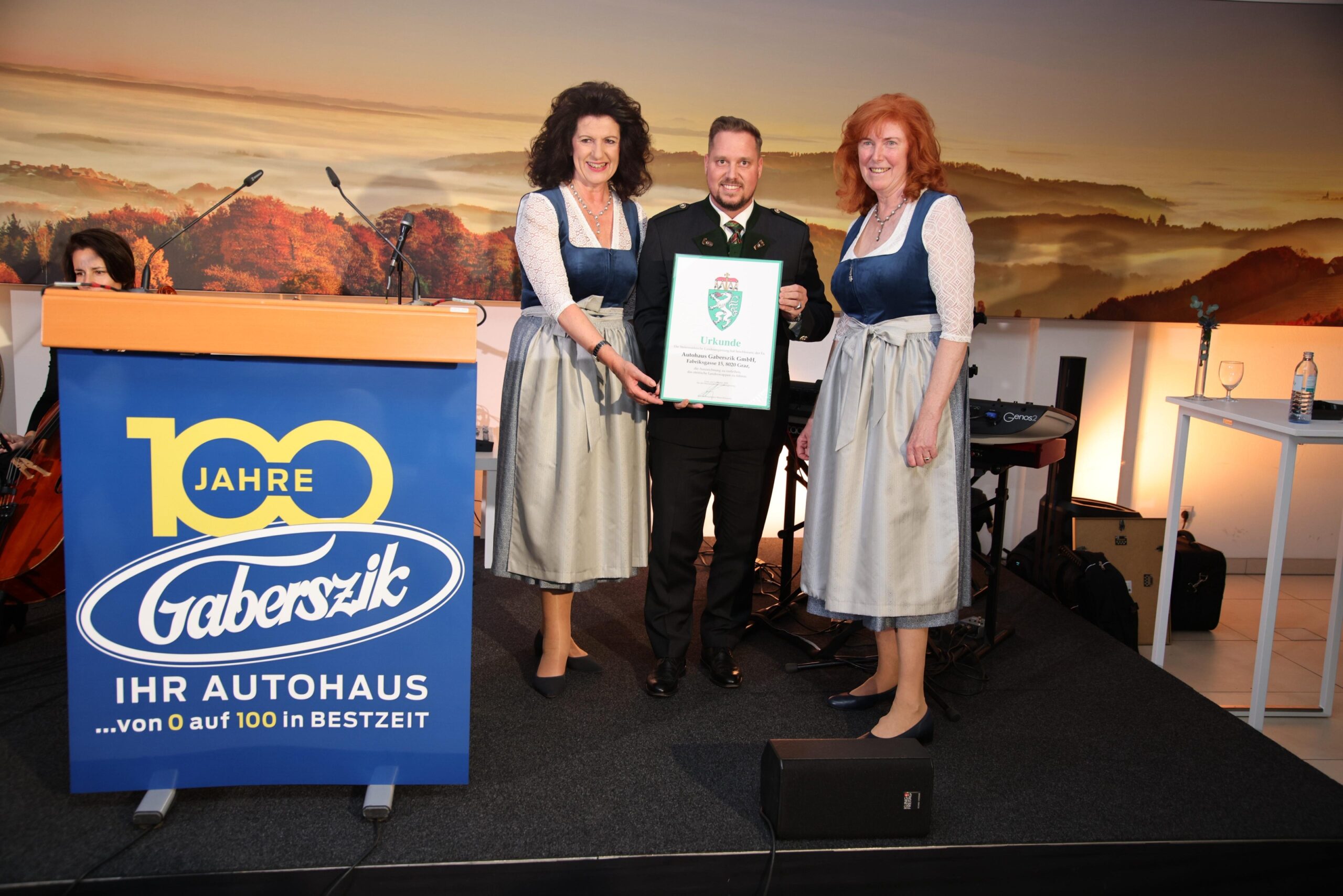 Drei Personen in bayerischer Tracht stehen auf einer Bühne bei den Holz steirischen Tischlertagen; der Mann in der Mitte hält eine Urkunde. Ein Podium mit der Aufschrift "100 Jahre Gaberszik" und ein landschaftlicher Hintergrund sind zu sehen.