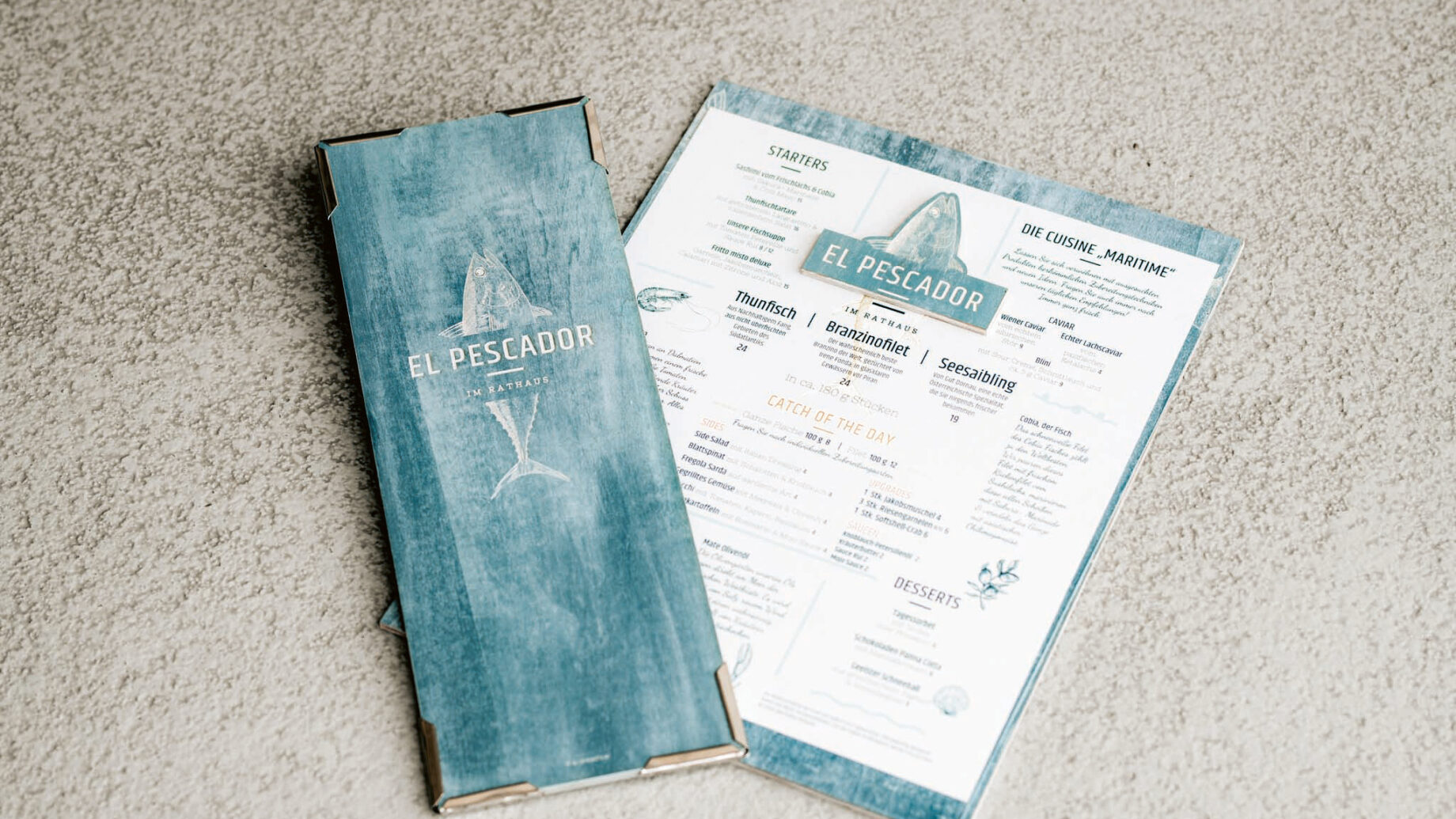 Eine blau-weiße Speisekarte für ein Fischrestaurant mit der Aufschrift "El Pescador" ist neben dem dazugehörigen blauen Umschlag abgebildet, die beide auf einer hellen, strukturierten Oberfläche liegen.