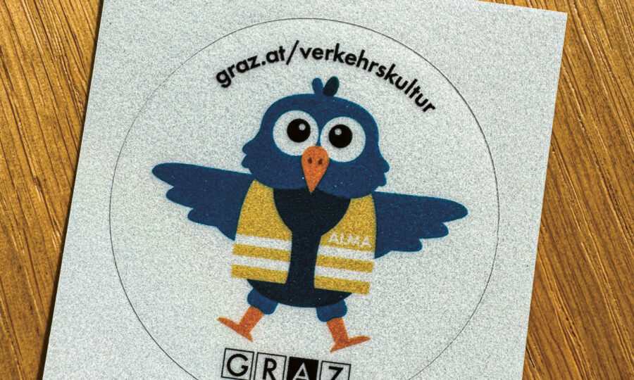 Ein Aufkleber zeigt einen blauen Cartoon-Vogel, der eine gelbe Sicherheitsweste mit der Aufschrift "ALMA" trägt. Über dem Vogel steht die URL "graz.at/verkehrskultur", darunter steht das Wort "GRAZ". Der Aufkleber befindet sich auf einer Holzfläche.