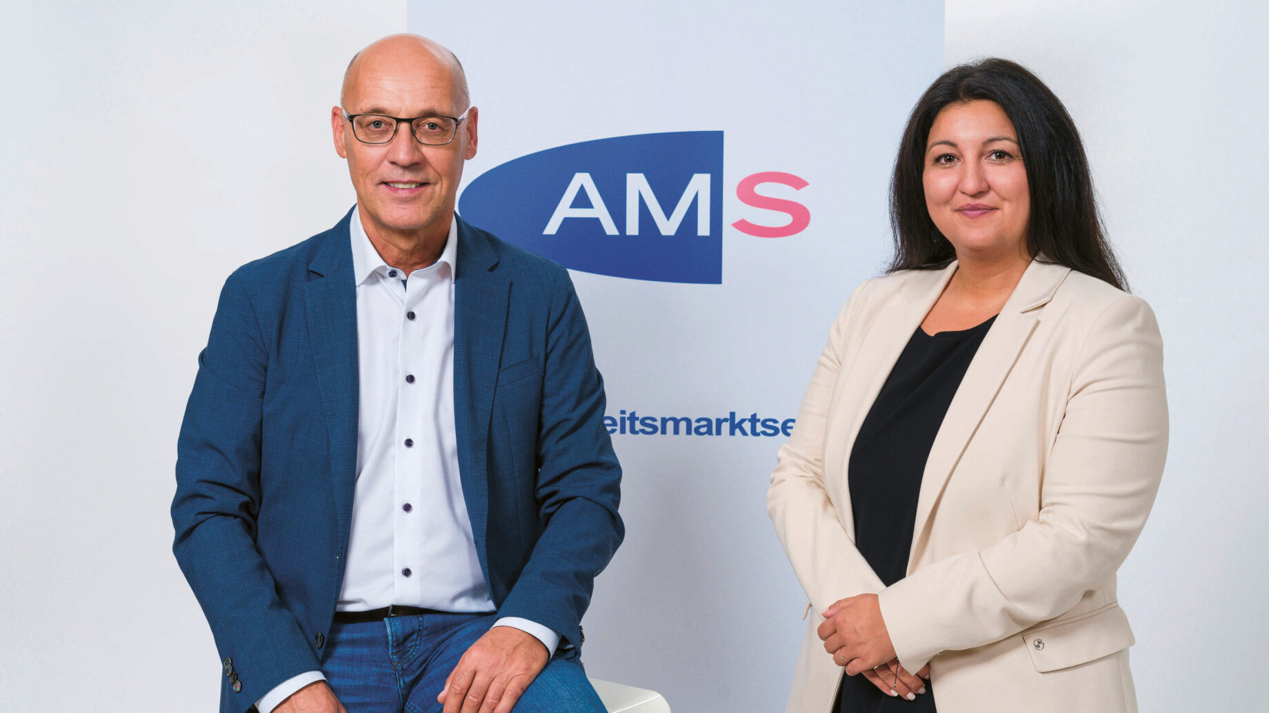 Zwei Personen in Geschäftskleidung stehen vor einem Hintergrund mit dem "AMS"-Logo. Der Mann auf der linken Seite sitzt auf einem Hocker und trägt einen blauen Anzug, die Frau auf der rechten Seite steht und trägt einen beigen Blazer. Beide lächeln.