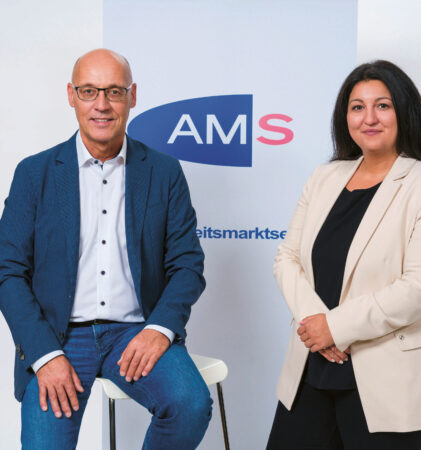 Zwei Personen in Geschäftskleidung stehen vor einem Hintergrund mit dem "AMS"-Logo. Der Mann auf der linken Seite sitzt auf einem Hocker und trägt einen blauen Anzug, die Frau auf der rechten Seite steht und trägt einen beigen Blazer. Beide lächeln.