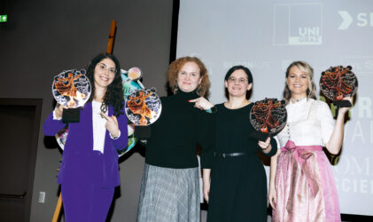 Vier Frauen stehen in einem Innenraum nebeneinander, lächeln und halten runde, bunte Auszeichnungen in der Hand. Sie sind formell bis halbformell gekleidet und posieren vor einem Bildschirm, auf dem Logos und Texte zu sehen sind.