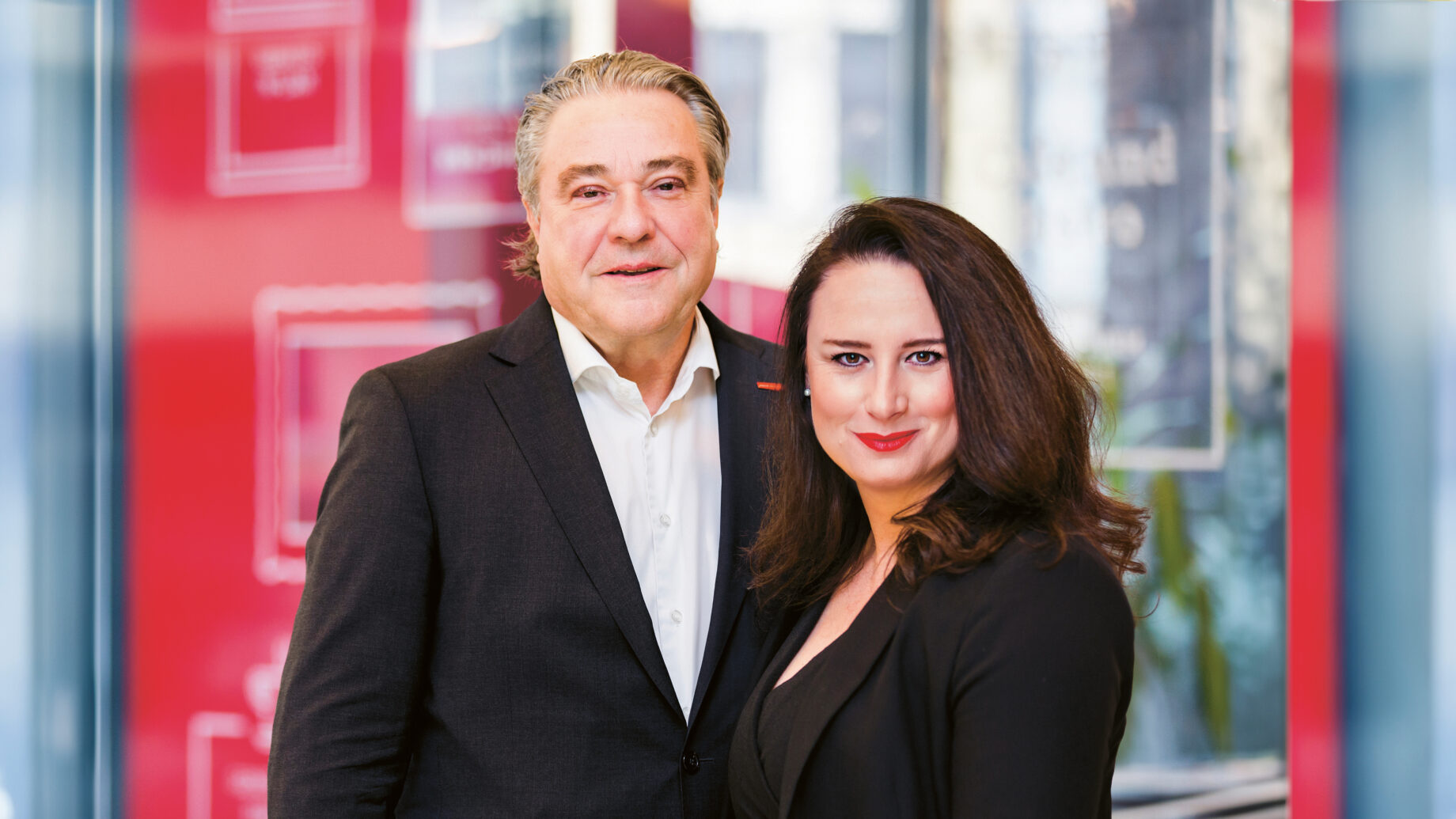 Ein Mann und eine Frau, beide in schwarzen Anzügen, stehen eng beieinander und lächeln in die Kamera vor einem modernen Bürohintergrund aus Rot und Glas.