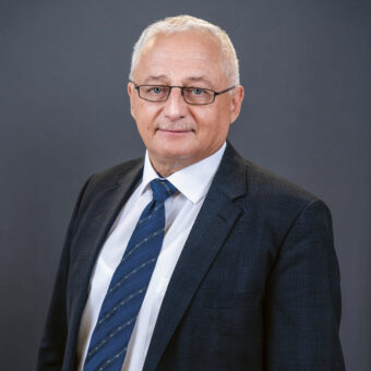 Ein älterer Mann mit kurzen grauen Haaren, Brille und Schnurrbart trägt einen dunklen Anzug, ein weißes Hemd und eine blau gemusterte Krawatte und steht vor einem schlichten dunkelgrauen Hintergrund.