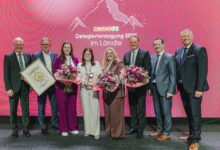 Acht Personen in Business-Kleidung posieren auf der Bühne mit Blumen, einer Trophäe und einer gerahmten Urkunde und feiern auf der "SPAR Delegiertentagung 2023 im Ländle", wo sich der Holzcluster Steiermark für eine nachhaltige Zukunft einsetzt.