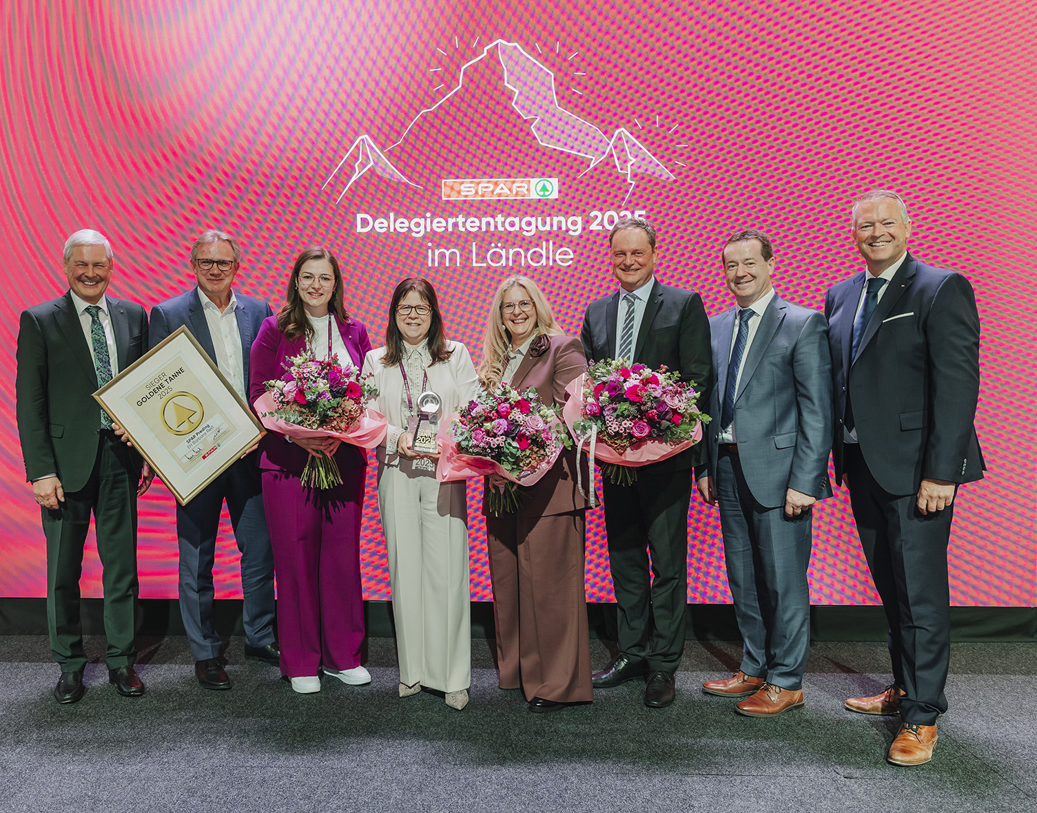 Acht Personen in Business-Kleidung posieren auf der Bühne mit Blumen, einer Trophäe und einer gerahmten Urkunde und feiern auf der "SPAR Delegiertentagung 2023 im Ländle", wo sich der Holzcluster Steiermark für eine nachhaltige Zukunft einsetzt.