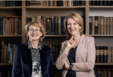 Zwei Frauen stehen lächelnd vor einem Bücherregal im Kompetenzzentrum der WKO Steiermark. Die eine trägt eine Brille, eine gemusterte Bluse und einen dunklen Blazer, die andere einen hellen Blazer, ein dunkles Oberteil und kurze blonde Haare.