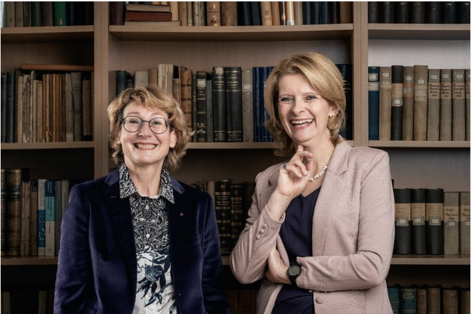 Zwei Frauen stehen lächelnd vor einem Bücherregal im Kompetenzzentrum der WKO Steiermark. Die eine trägt eine Brille, eine gemusterte Bluse und einen dunklen Blazer, die andere einen hellen Blazer, ein dunkles Oberteil und kurze blonde Haare.