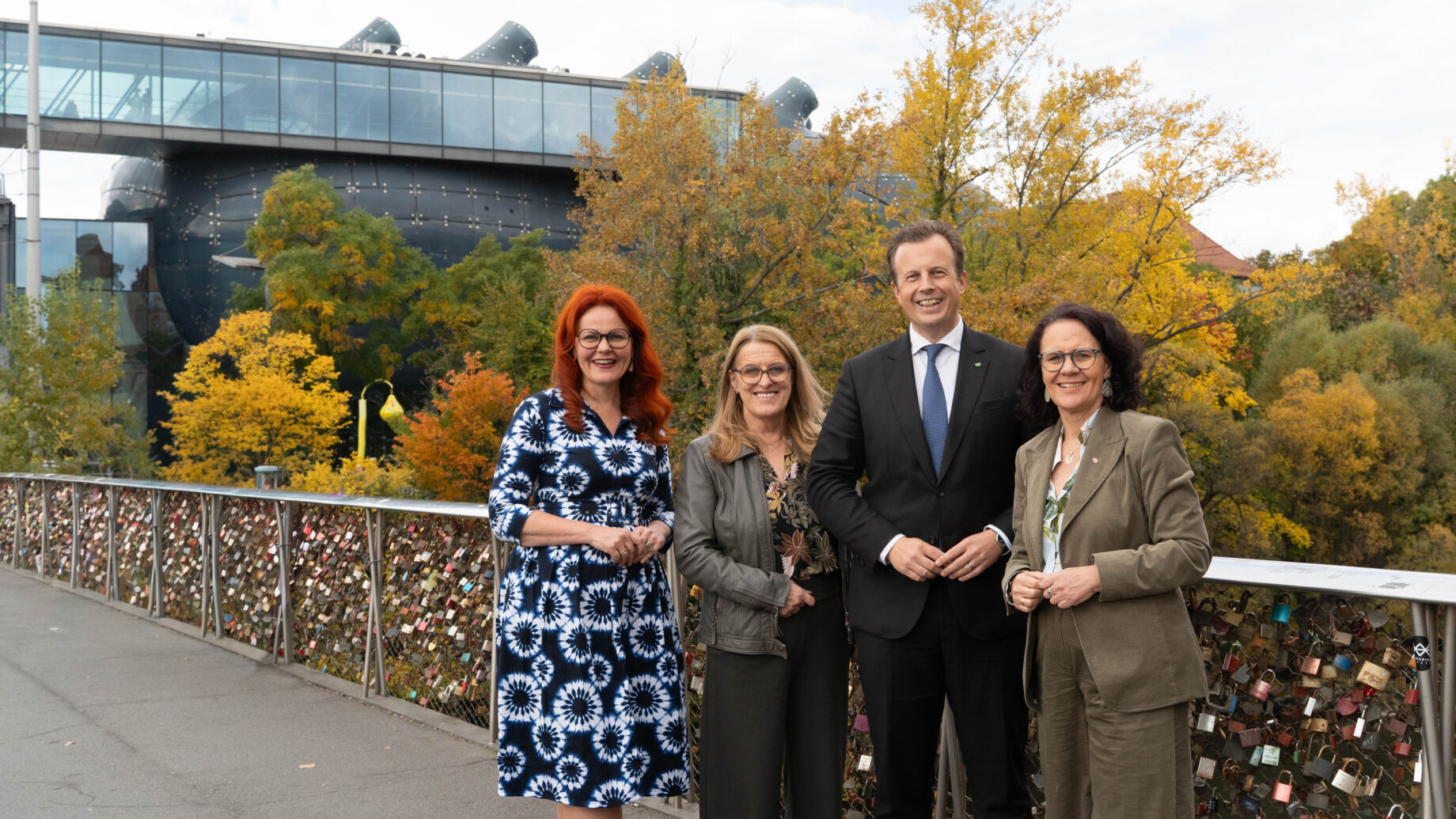 Vier Erwachsene, drei Frauen und ein Mann, stehen lächelnd auf einer mit Liebesschlössern geschmückten Brücke. Im Hintergrund sind Herbstbäume und das moderne Glasgebäude der Merkur Versicherung zu sehen, auf dem das steirische Landeswappen abgebildet ist.