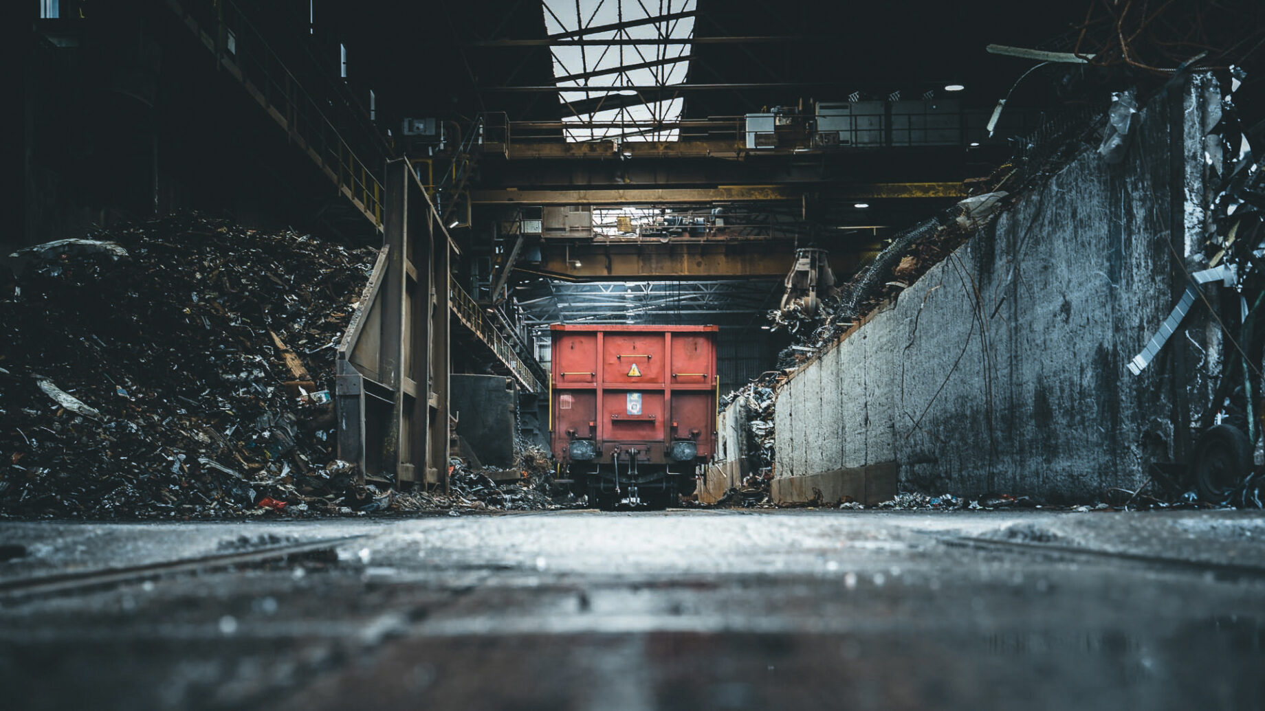 Ein roter Industrietriebwagen steht in einer dunklen, unübersichtlichen Lagerhalle voller Metallschrotthaufen und Schutt unter einem teilweise offenen Dach, das natürliches Licht hereinlässt.