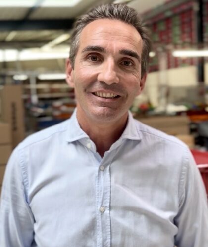 Ein Mann mit kurzen braunen Haaren, der ein hellblaues Button-up-Hemd trägt, lächelt in die Kamera in einem gut beleuchteten Innenraum mit Regalen und Kisten im Hintergrund - perfekt für "3 Fragen an MCG-Vorstand Martin Ullrich".