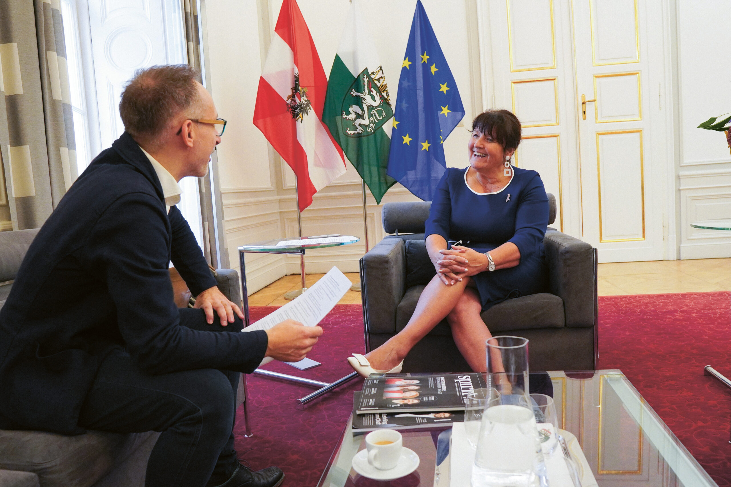 Ein Mann interviewt eine lächelnde Frau, die in einem Büro sitzt, hinter dem österreichische, regionale und EU-Fahnen wehen. Sie sitzen auf Sesseln neben einem Glastisch mit Zeitschriften, Papieren, einer Wasserkaraffe und einer Kaffeetasse.