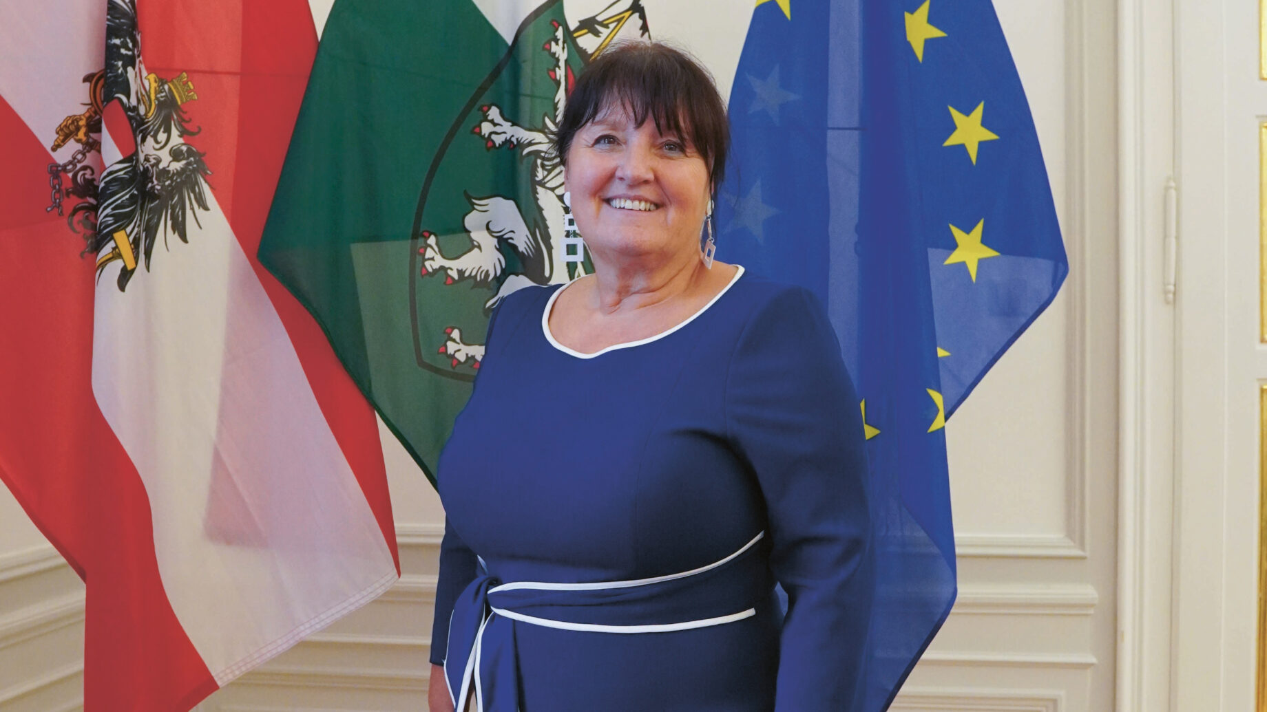Eine Frau in einem blauen Kleid steht lächelnd vor drei Fahnen: Österreich, Steiermark und die Europäische Union. Sie steht in einem Innenraum auf einem Parkettboden mit weiß getäfelten Wänden.