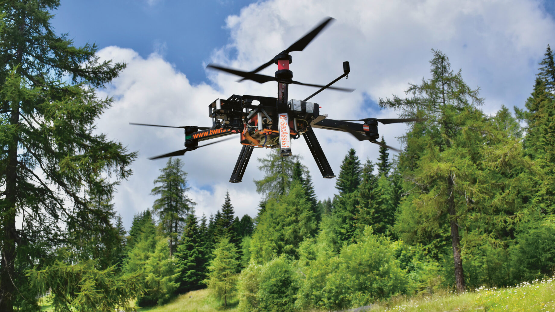 Eine schwarze Drohne mit vier Rotoren fliegt über einen Feldweg in einem grünen Wald, umgeben von hohen Bäumen und unter einem teilweise bewölkten blauen Himmel.