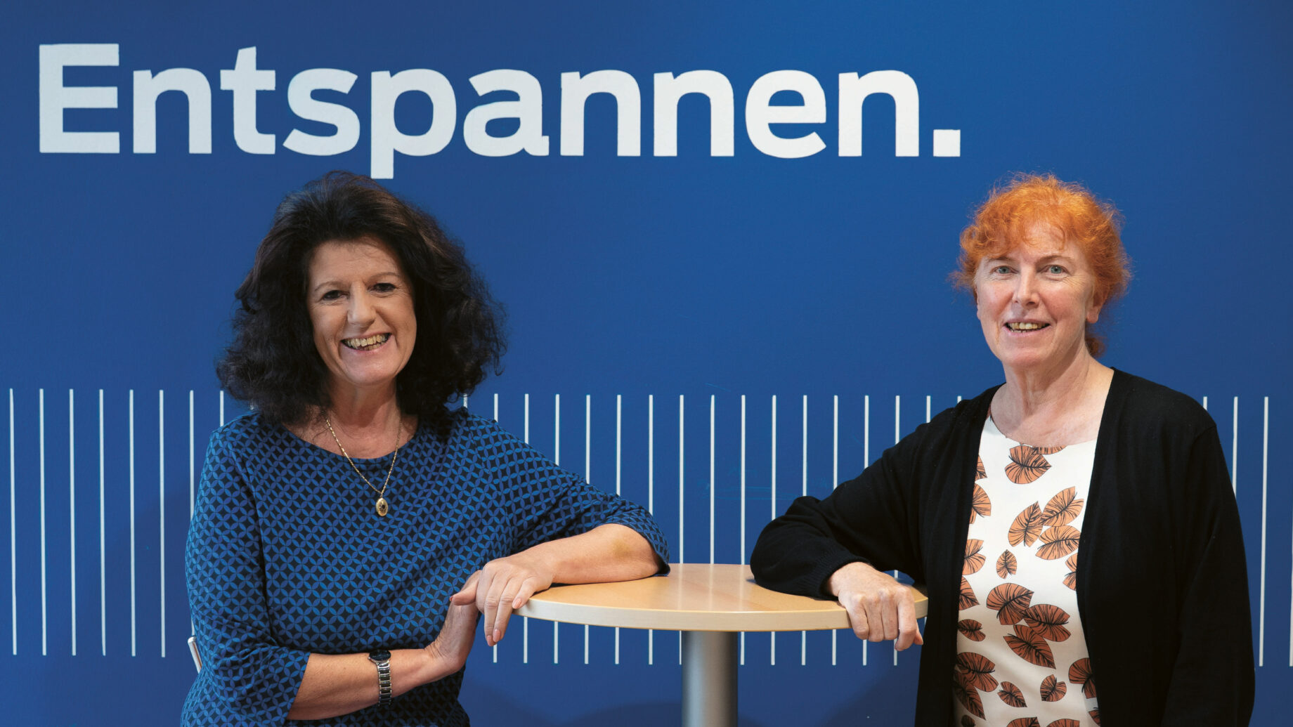 Zwei Frauen sitzen auf Hockern an einem hohen runden Tisch und lächeln vor einer blauen Wand, über der in großen weißen Buchstaben das Wort "Entspannen." steht.