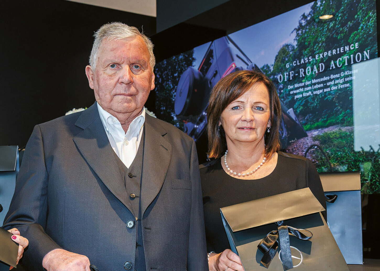 Drei ältere Menschen stehen in einem Haus und halten jeweils eine schwarze Mercedes-Benz Geschenktüte in der Hand. Sie sind förmlich gekleidet und posieren für die Kamera. Im Hintergrund ist ein Werbedisplay von Mercedes-Benz zu sehen.