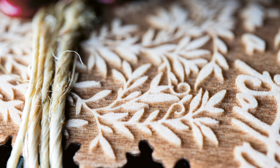 Nahaufnahme eines strukturierten braunen Stoffes mit einem komplizierten, erhabenen Blattmuster und mehreren Strängen beiger Schnüre, die darüber liegen. Der Hintergrund ist sanft verschwommen.