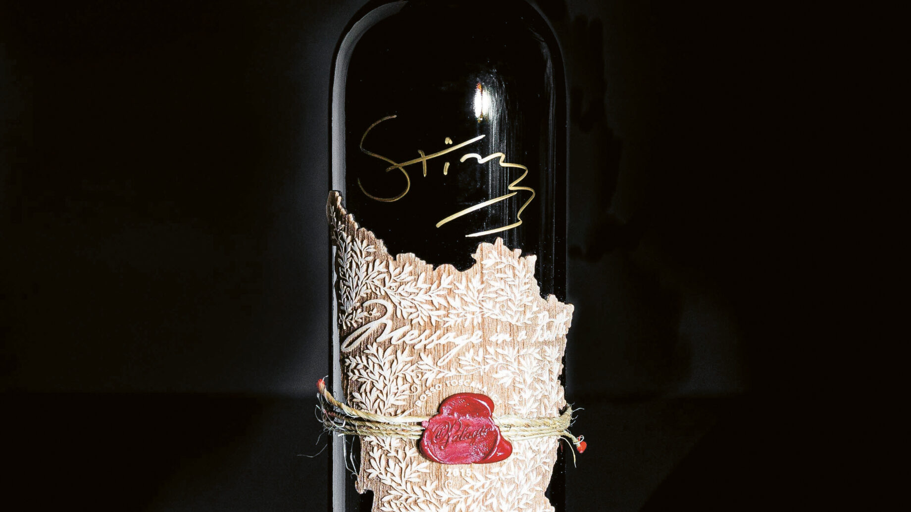 Eine dunkle Weinflasche mit einem roten Wachssiegel auf der Oberseite und goldener Handschrift auf dem Glas. Um die Flasche ist ein strukturiertes, metallisch anmutendes Etikett mit einer gebundenen Schnur und einem roten Wachsstempel gewickelt. Die Flasche ist vor einem schwarzen Hintergrund platziert.
