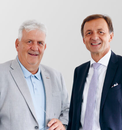 Zwei formell gekleidete Männer mittleren Alters stehen nebeneinander und lächeln. Der eine trägt einen hellgrauen Anzug mit einem blauen Hemd, der andere einen dunklen Anzug mit weißem Hemd und Krawatte. Der Hintergrund ist schlicht und hell.