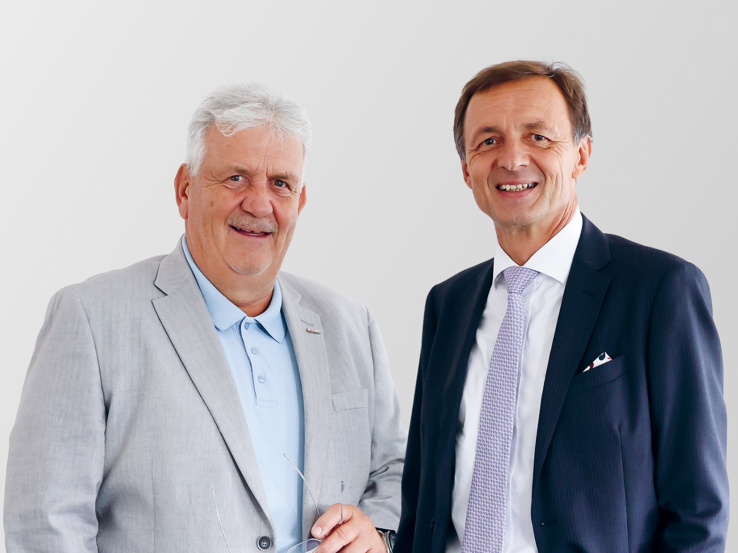 Zwei formell gekleidete Männer mittleren Alters stehen nebeneinander und lächeln. Der eine trägt einen hellgrauen Anzug mit einem blauen Hemd, der andere einen dunklen Anzug mit weißem Hemd und Krawatte. Der Hintergrund ist schlicht und hell.