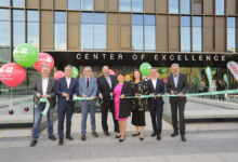 Acht Personen in Geschäftskleidung stehen vor einem modernen Gebäude mit der Aufschrift "Center of Excellence" im Siemens Energy Werk Wollsdorf und halten ein grünes Band für eine Zeremonie zum Durchschneiden des Bandes. Im Hintergrund sind bunte Luftballons und große Fenster zu sehen.