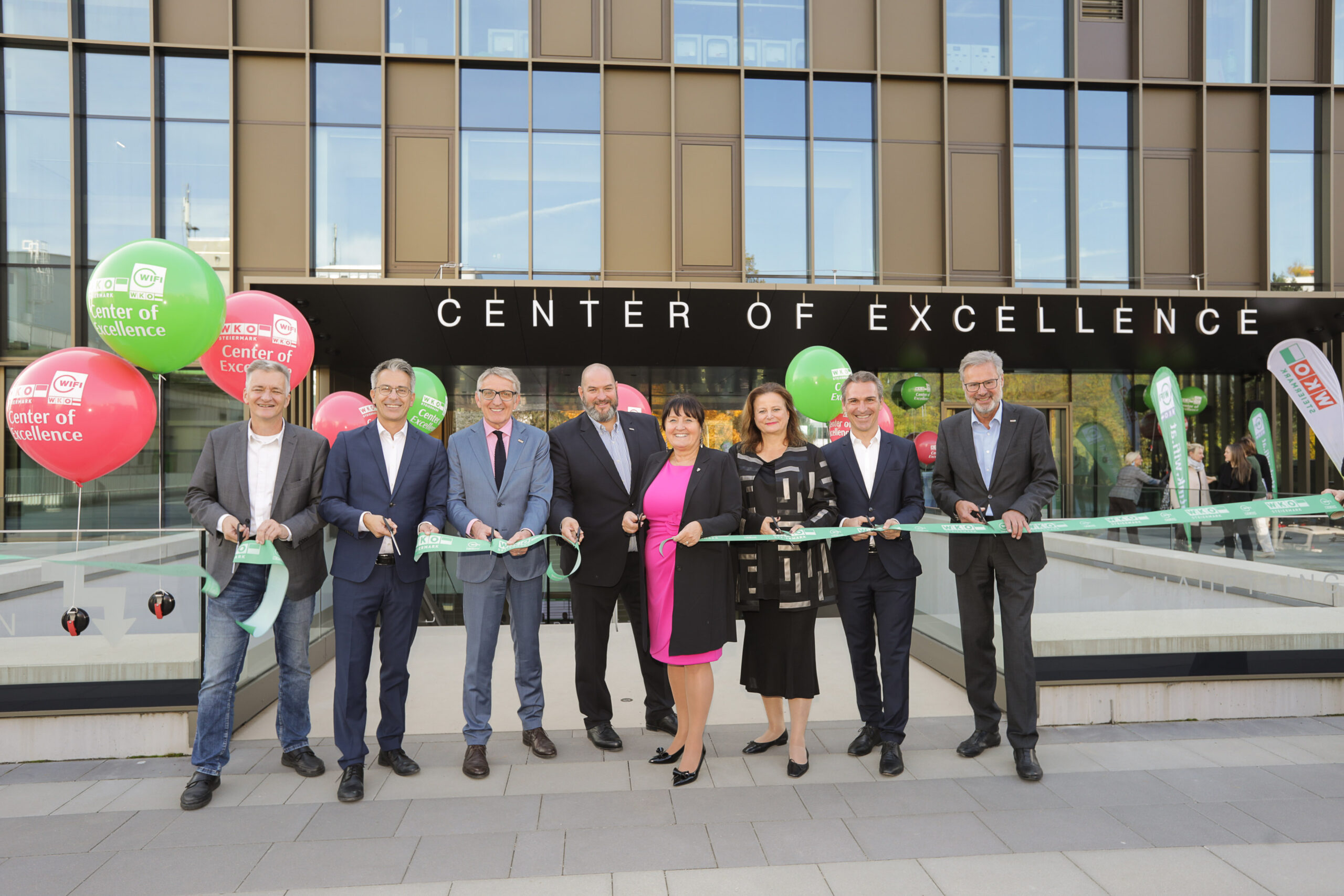 Acht Personen in Geschäftskleidung stehen vor einem modernen Gebäude mit der Aufschrift "Center of Excellence" im Siemens Energy Werk Wollsdorf und halten ein grünes Band für eine Zeremonie zum Durchschneiden des Bandes. Im Hintergrund sind bunte Luftballons und große Fenster zu sehen.