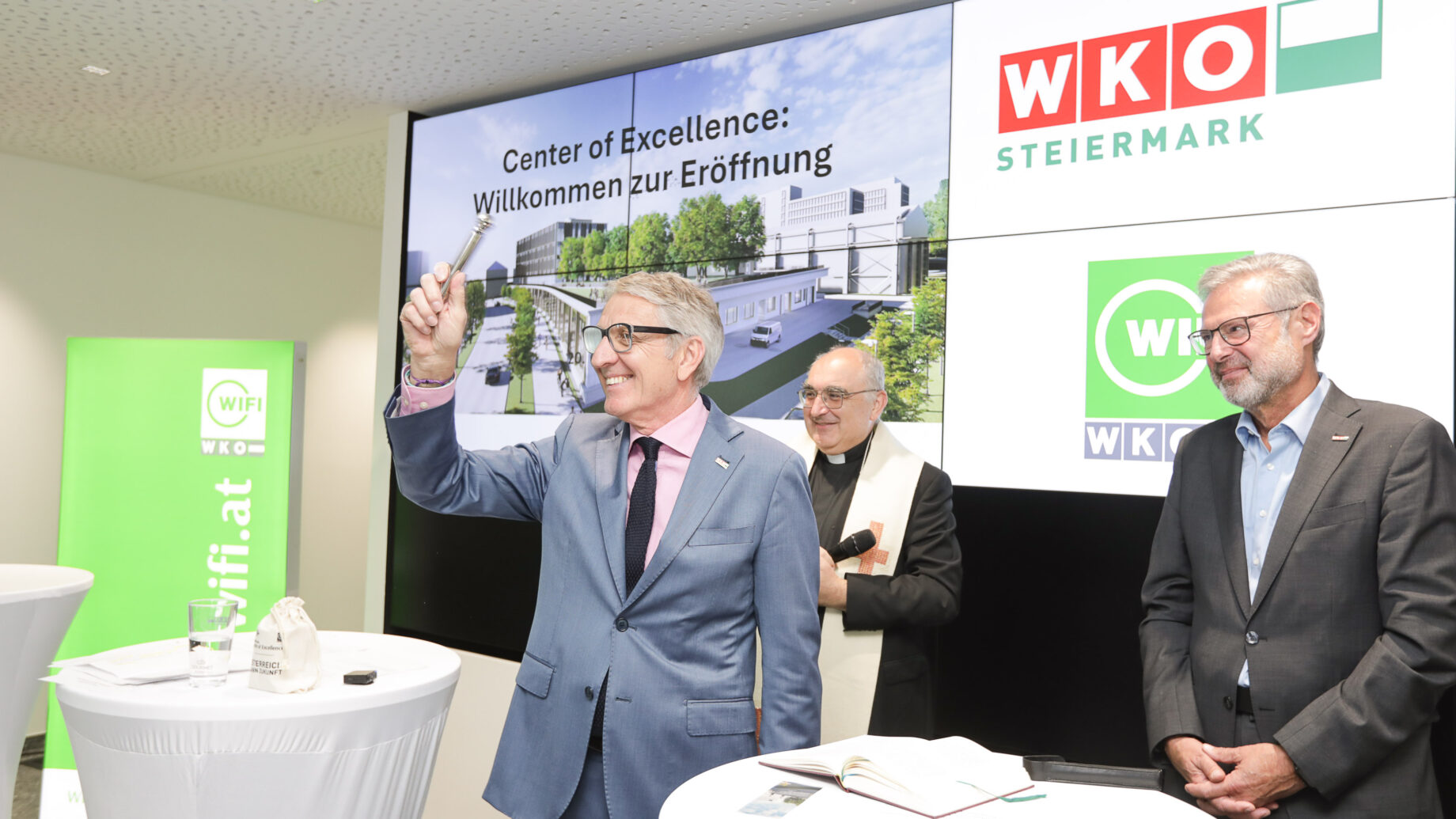 Drei Männer stehen an einem Podium vor einem großen Bildschirm, auf dem "Center of Excellence: Willkommen zur Eröffnung" und das Logo der WKO Steiermark. Ein Mann hält lächelnd einen Schlüssel hoch und feiert die Eröffnung des Siemens Energy Werk Wollsdorf.