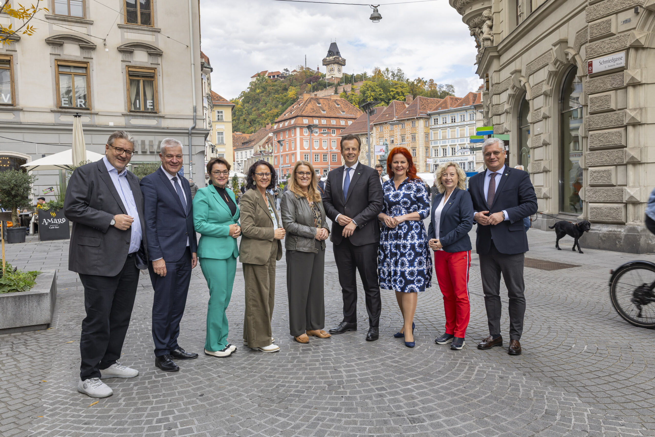 Eine Gruppe von neun Erwachsenen in Geschäftskleidung posiert und lächelt auf einer kopfsteingepflasterten Straße in einer europäischen Stadt, mit historischen Gebäuden, dem steirischen Landeswappen und einem Hügel mit einem Uhrenturm im Hintergrund - und fängt damit den Geist der Merkur Versicherung ein.