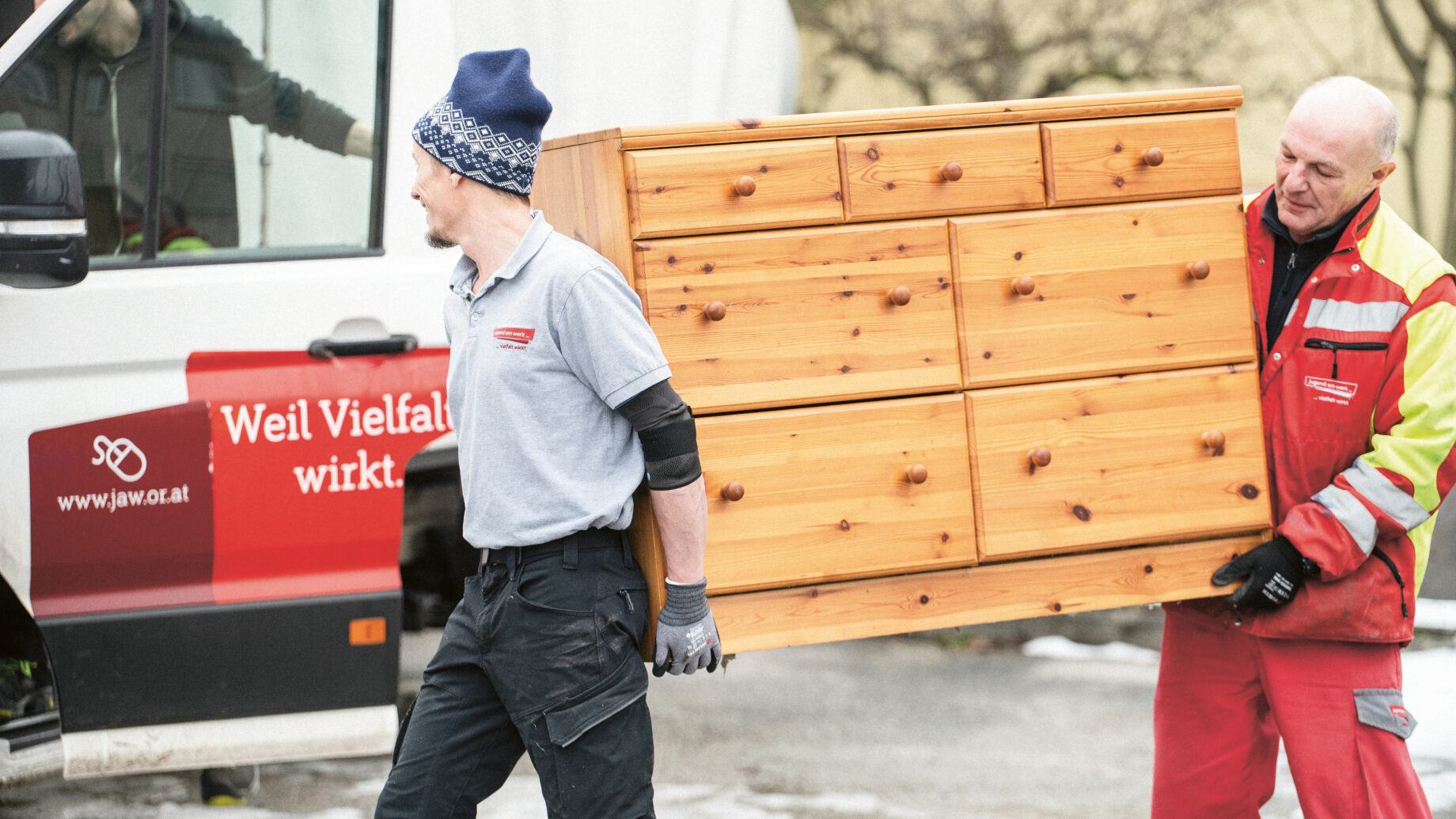 Zwei Männer, einer in grauer und blauer Arbeitskleidung, der andere in rotem Overall, tragen eine große Holzkommode nach draußen, neben einen weiß-roten Lieferwagen, auf leicht verschneitem Boden.