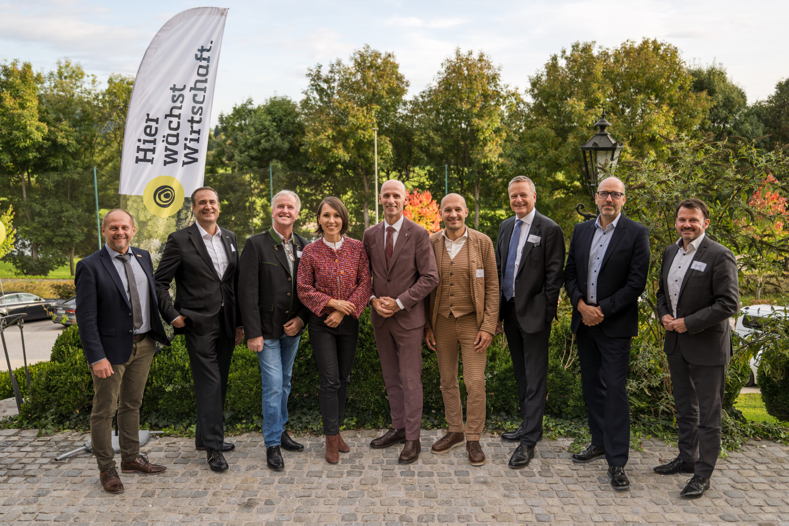 Eine Gruppe von zehn Personen, gekleidet in Business- oder eleganter Freizeitkleidung, steht im Freien auf einem Kopfsteinpflasterweg vor einer Grünanlage. Eine große Fahne mit dem Logo des Autohauses Gaberszik und deutschem Text ist zu sehen, die 100 Jahre Autohaus Gaberszik markiert.
