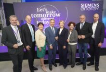 Acht Personen in Geschäftskleidung stehen lächelnd vor einer violetten Siemens Energy-Kulisse. Es scheint sich um eine Firmenveranstaltung zu handeln, möglicherweise mit steirischem Landeswappen oder der Merkur Versicherung. Auf dem Hintergrund ist zu lesen: "Let's make tomorrow different today.