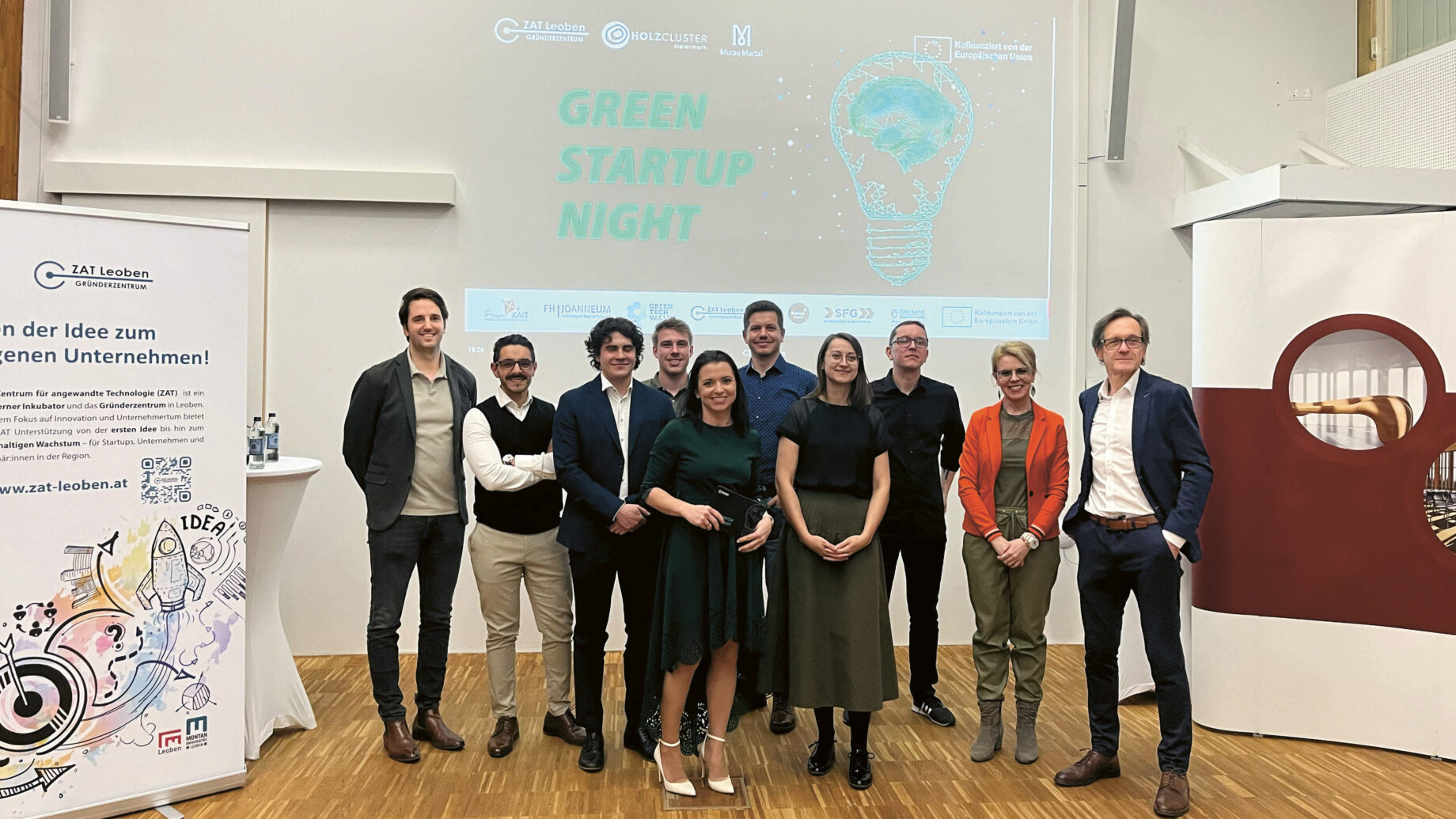Neun Personen stehen in einem Innenraum zusammen und lächeln in die Kamera vor einem Bildschirm mit der Aufschrift "Green Startup Night" und einer Glühbirnengrafik. Die Umgebung scheint eine professionelle Veranstaltung oder Konferenz zu sein.