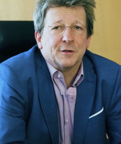 Ein Mann mittleren Alters mit Brille und zerzaustem Haar, der eine blaue Anzugsjacke über einem hellvioletten Hemd trägt, sitzt in einem Bürostuhl vor einem hölzernen Hintergrund.