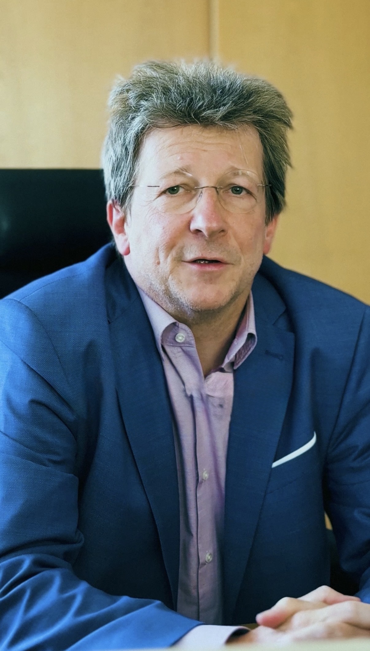 Ein Mann mittleren Alters mit Brille und zerzaustem Haar, der eine blaue Anzugsjacke über einem hellvioletten Hemd trägt, sitzt in einem Bürostuhl vor einem hölzernen Hintergrund.