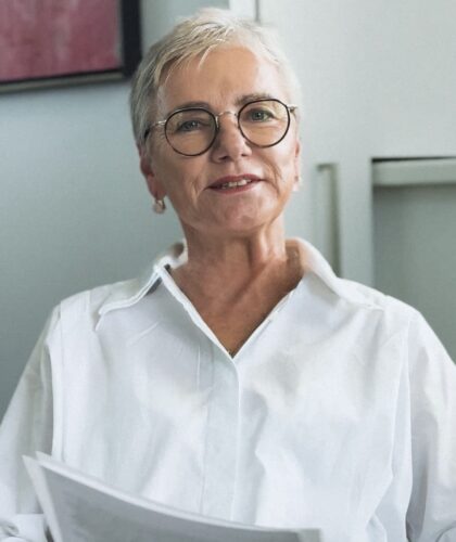Eine ältere Frau mit kurzen grauen Haaren und Brille, die ein weißes Hemd trägt, hält Papiere in der Hand und lächelt. Sie sitzt drinnen in einem gut beleuchteten Raum.