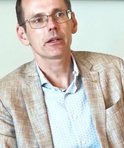 Ein Mann mit kurzen braunen Haaren und Brille, der einen hellbraun karierten Blazer und ein gemustertes weißes Hemd trägt.