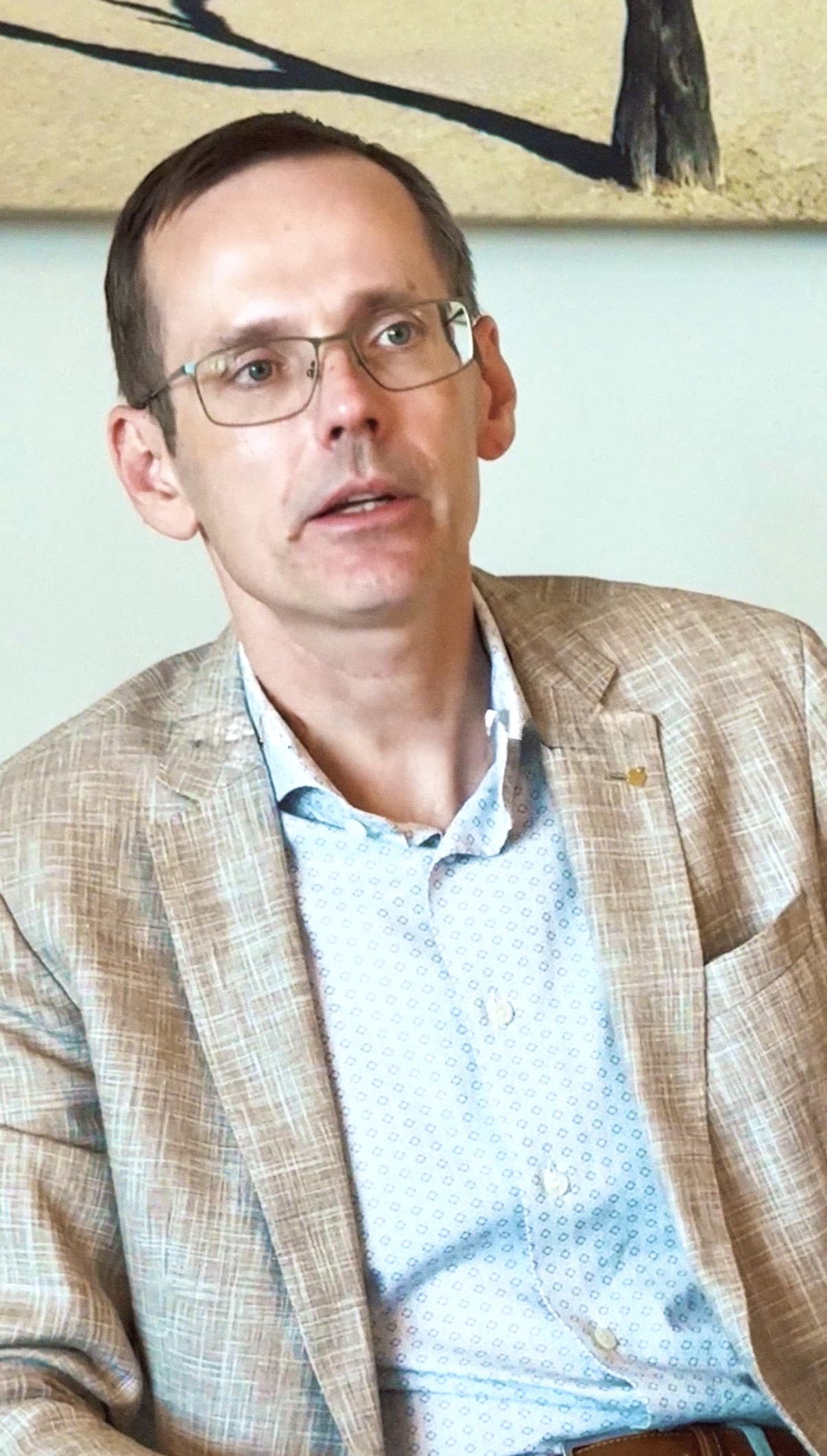 Ein Mann mit kurzen braunen Haaren und Brille, der einen hellbraun karierten Blazer und ein gemustertes weißes Hemd trägt.