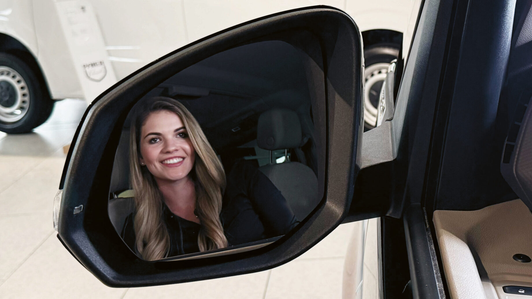 Eine Frau mit langen blonden Haaren lächelt und spiegelt sich im Seitenspiegel eines Autos in einem Ausstellungsraum, in dem im Hintergrund weiße Fahrzeuge zu sehen sind.