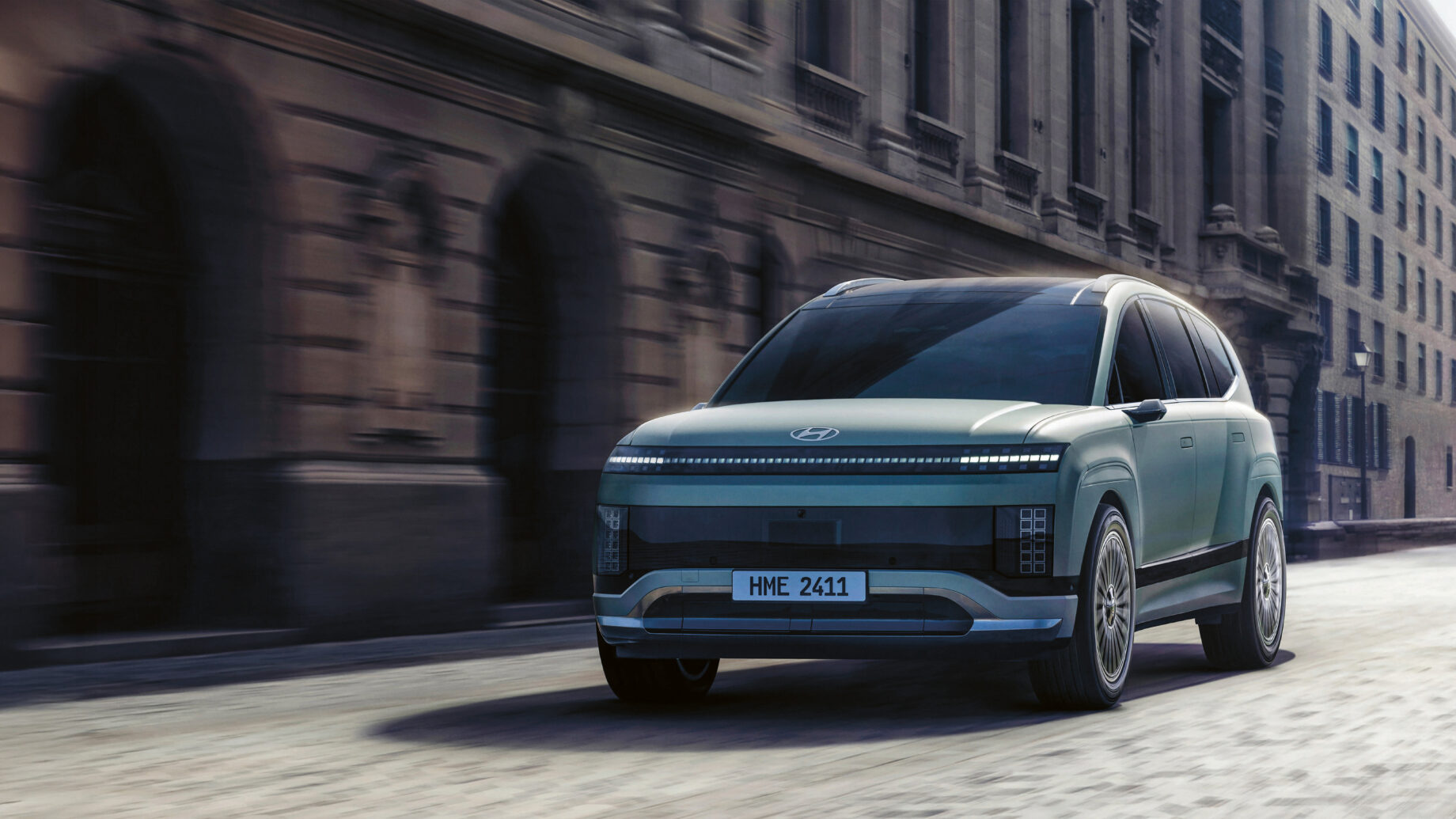 Ein futuristischer silberner Geländewagen fährt an einem bewölkten Tag eine von alten, verschnörkelten Steingebäuden gesäumte Stadtstraße entlang.