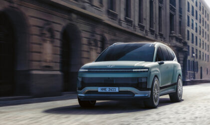 Ein futuristischer silberner Geländewagen fährt an einem bewölkten Tag eine von alten, verschnörkelten Steingebäuden gesäumte Stadtstraße entlang.