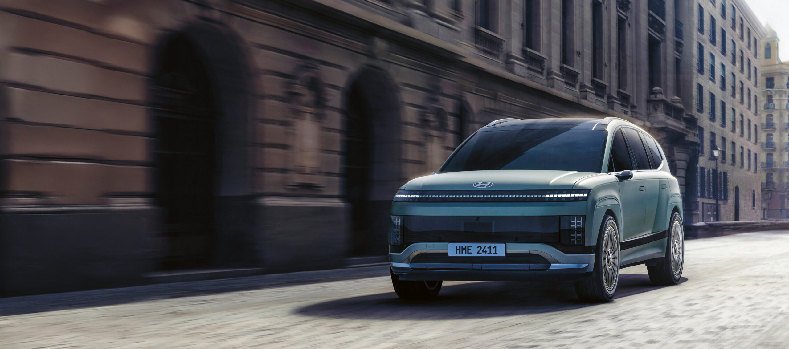 Ein futuristischer silberner Geländewagen fährt an einem bewölkten Tag eine von alten, verschnörkelten Steingebäuden gesäumte Stadtstraße entlang.