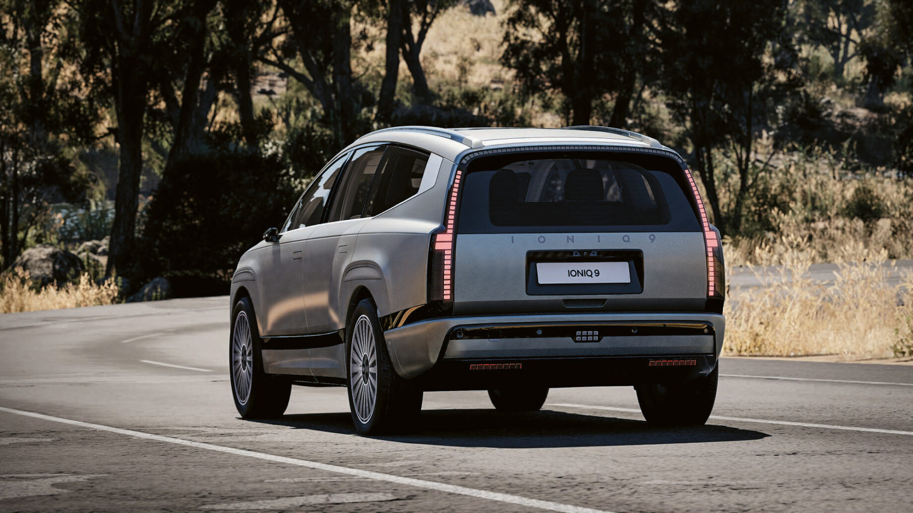 Ein silberner Hyundai Ioniq 7 Elektro-SUV fährt an einem sonnigen Tag auf einer gepflasterten Straße, umgeben von Bäumen und trockenem Gras, von hinten gesehen.