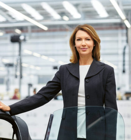 Eine Frau in einem Geschäftsanzug steht selbstbewusst neben einem Auto in einer hellen, modernen Industrie- oder Produktionsanlage mit großen Fenstern und Oberlichtern.