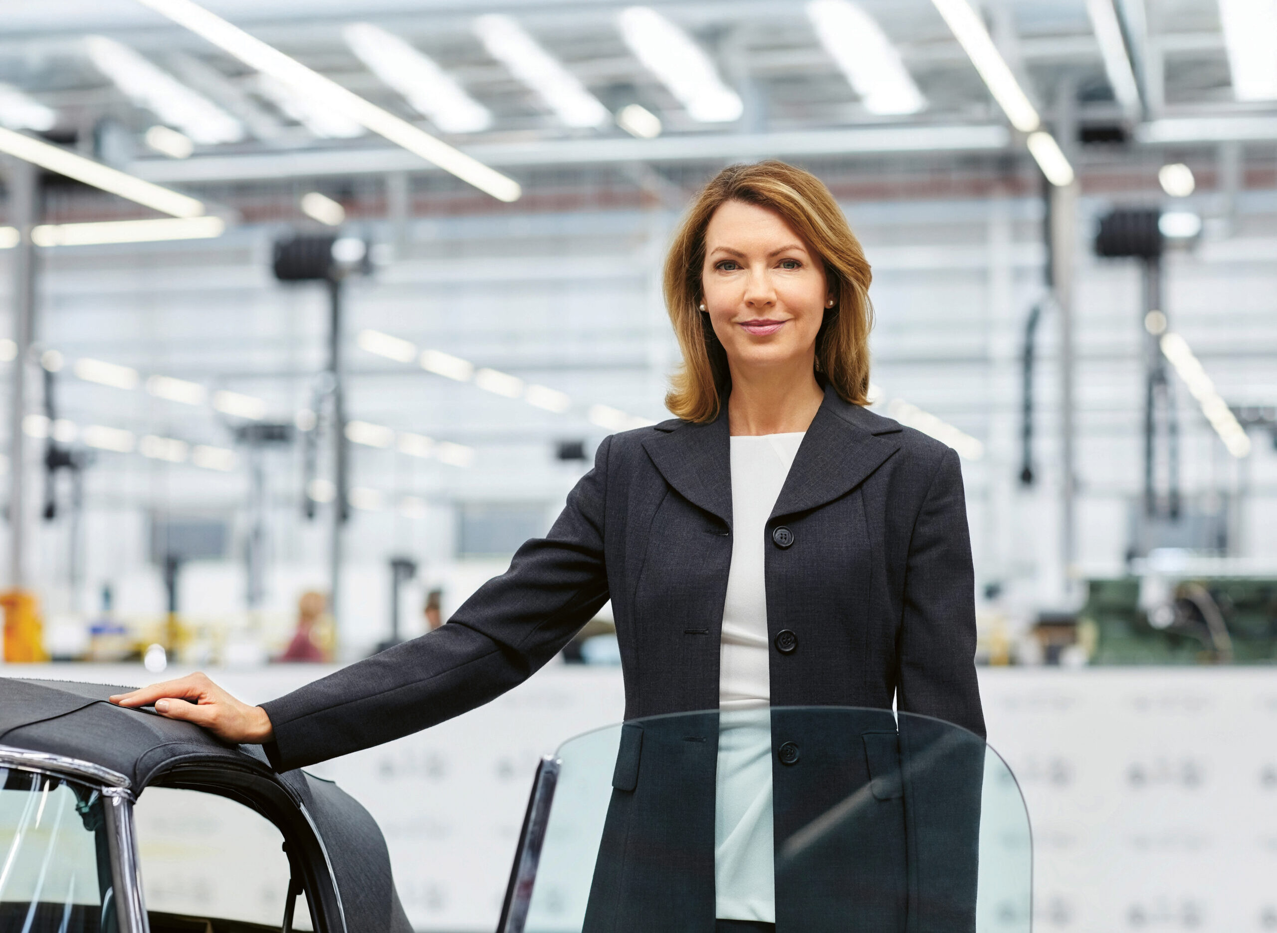 Eine Frau in einem Geschäftsanzug steht selbstbewusst neben einem Auto in einer hellen, modernen Industrie- oder Produktionsanlage mit großen Fenstern und Oberlichtern.