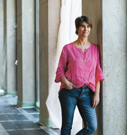 Eine Person mit kurzen dunklen Haaren, die ein hellrosa Hemd und eine blaue Jeans trägt, steht lässig an einem großen Fenster in einem gut beleuchteten Flur mit Betonwänden und unbearbeiteten Böden.