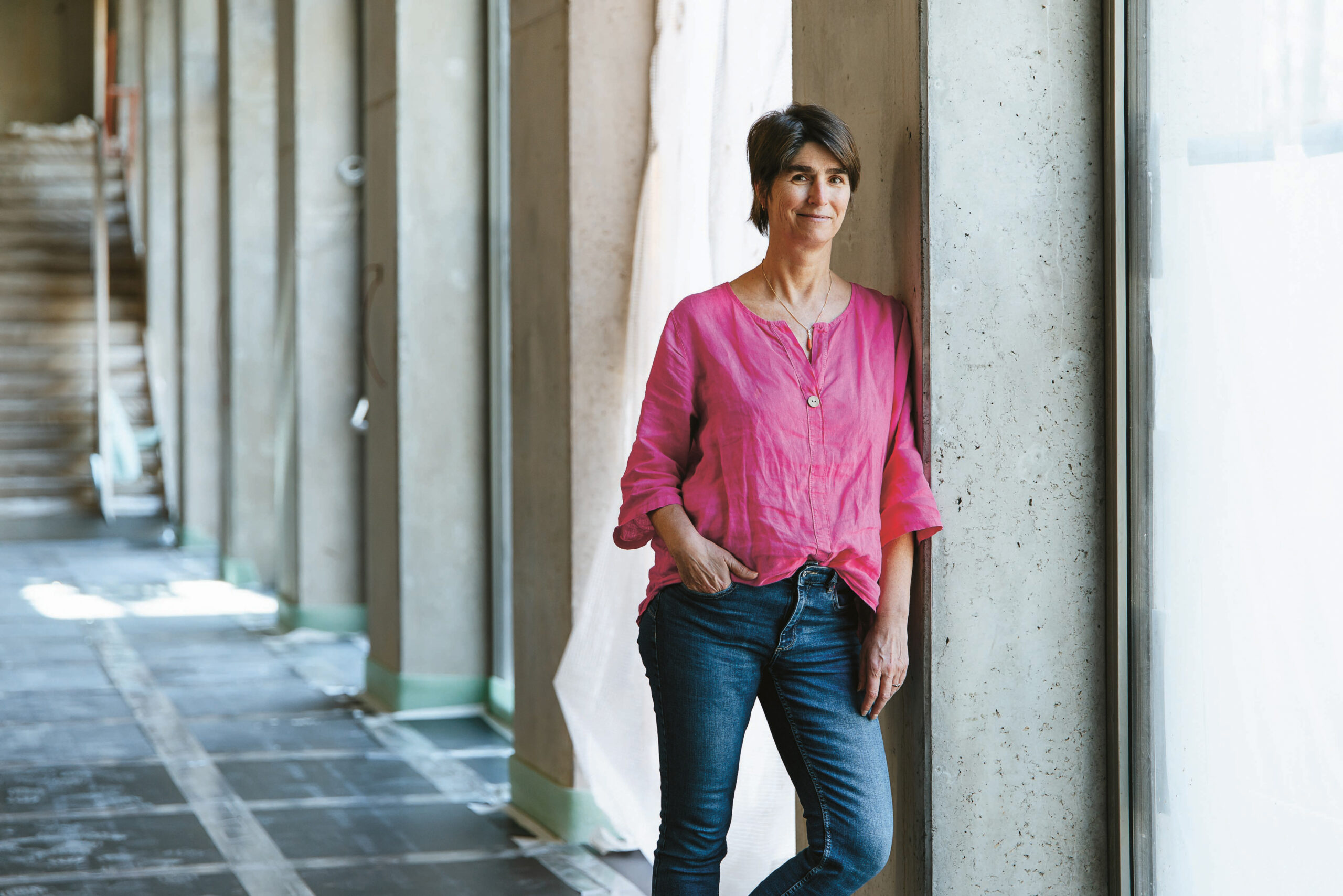 Eine Person mit kurzen dunklen Haaren, die ein hellrosa Hemd und eine blaue Jeans trägt, steht lässig an einem großen Fenster in einem gut beleuchteten Flur mit Betonwänden und unbearbeiteten Böden.
