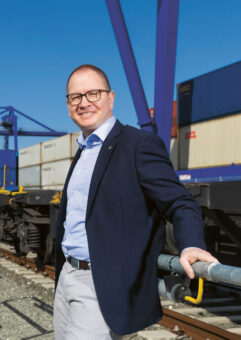 Ein Mann mit Brille, marineblauem Blazer und hellen Hosen steht an einem sonnigen Tag lächelnd neben einem Eisenbahngleis mit gestapelten Frachtcontainern und großen Kränen im Hintergrund.