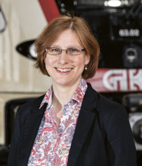 Eine Frau mit Brille und kurzen braunen Haaren, die eine gemusterte Bluse und einen dunklen Blazer trägt, steht lächelnd vor einem modernen Zug.