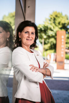 Eine Frau in weißem Blazer und rotem Kleid steht mit verschränkten Armen lächelnd neben einer spiegelnden Glaswand im Freien; im Hintergrund sind Bäume und Gebäude zu sehen.